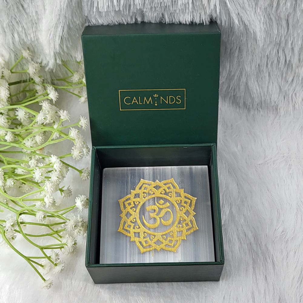 Gold Om symbol selenite plate in a green calminds box with white flowers on a gray background