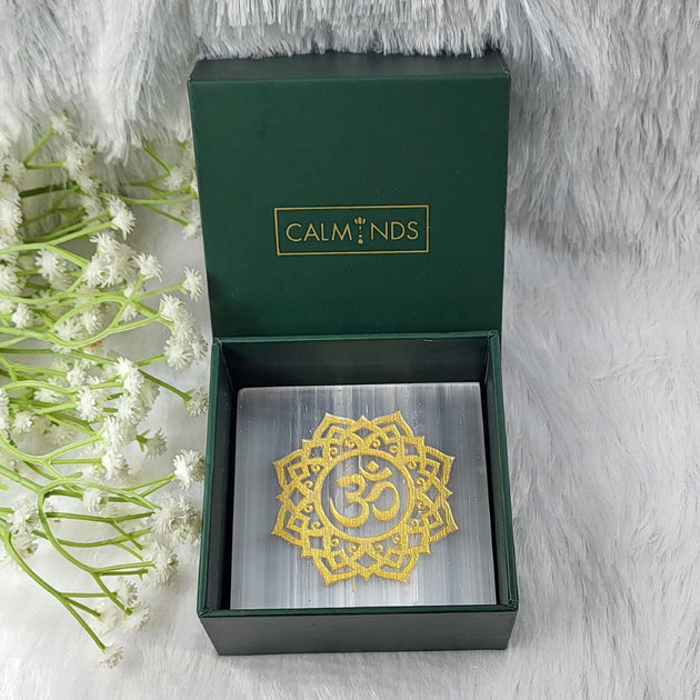 Gold Om symbol selenite plate in a green calminds box with white flowers on a gray background