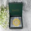 Gold Om symbol selenite plate in a green calminds box with white flowers on a gray background