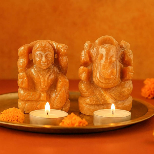 Two peach sunstone laksmi ganesh idol pair on a tray with two lit candles against an orange backgrounds