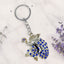 Keychain with peacock design and blue stones on a white background with lavender flowers.