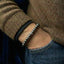 Close-up of a person wearing a beaded bracelet on a brown sweater and blue jeans.