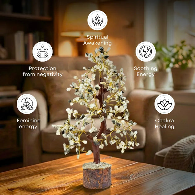A decorative crystal tree with white and gray stones and gold-toned accents on a wooden table, shown in a cozy living room setting with overlaid benefit icons and text: Spiritual Awakening, Protection from negativity, Soothing Energy, Feminine energy, and Chakra Healing.