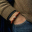 Close-up of a wrist wearing a red beaded bracelet with a brown sweater and blue jeans.