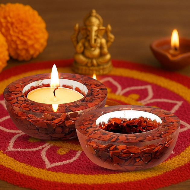Two lit candles in decorative red jasper holders on a colorful fabric background with a statue of Ganesha.