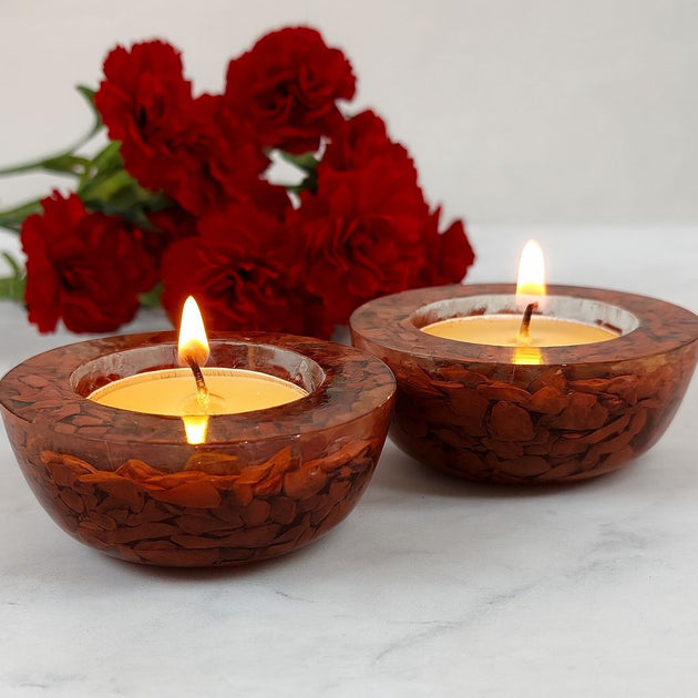 Two red candle holders with lit candles on a white surface, with red flowers in the background.