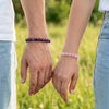 Two people holding hands with purple and pink beaded bracelets against a blurred green field background.