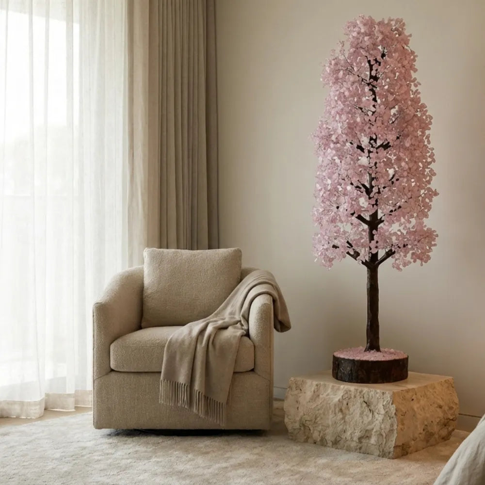 Decorative pink flowering tree on a stone base next to a beige armchair in a room with light-colored walls and curtains.