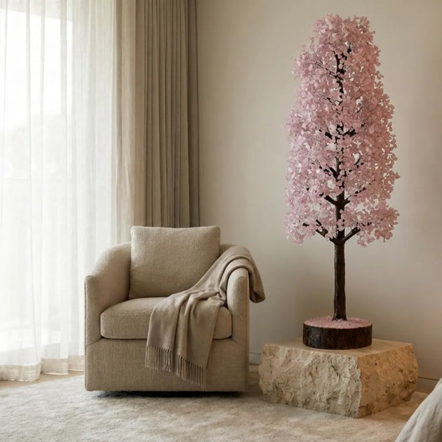 Decorative pink flowering tree on a stone base next to a beige armchair in a room with light-colored walls and curtains.