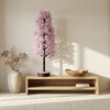Decorative indoor plant with pink blossoms on a wooden shelf in a room with a vase and bowl.