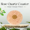 Rose Quartz Coaster with a gold emblem on a light gray background with greenery.