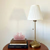 Table lamp with white shade and gold base on a wooden surface with books and a pink swan sculpture.