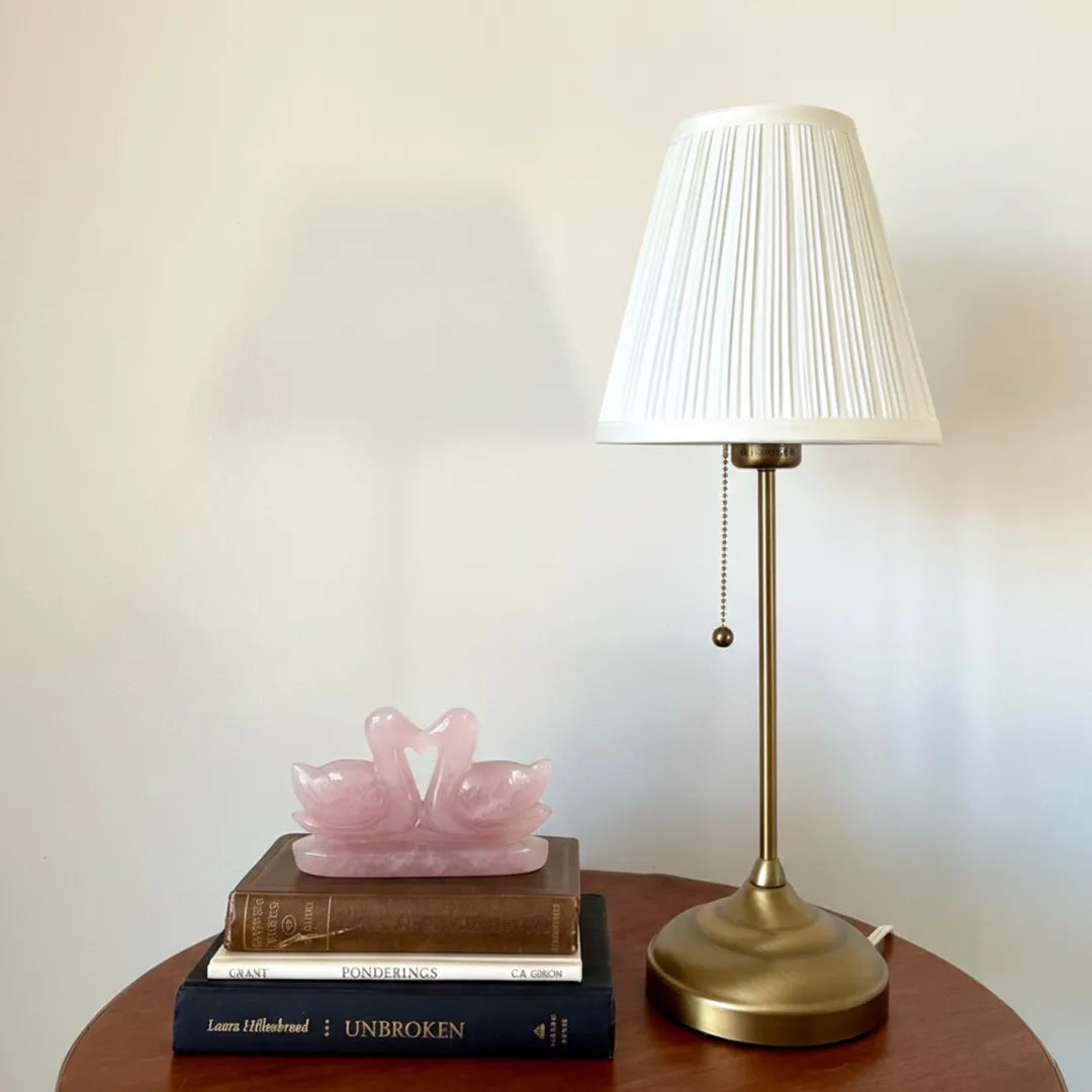 Table lamp with white shade and gold base on a wooden surface with books and a pink swan sculpture.