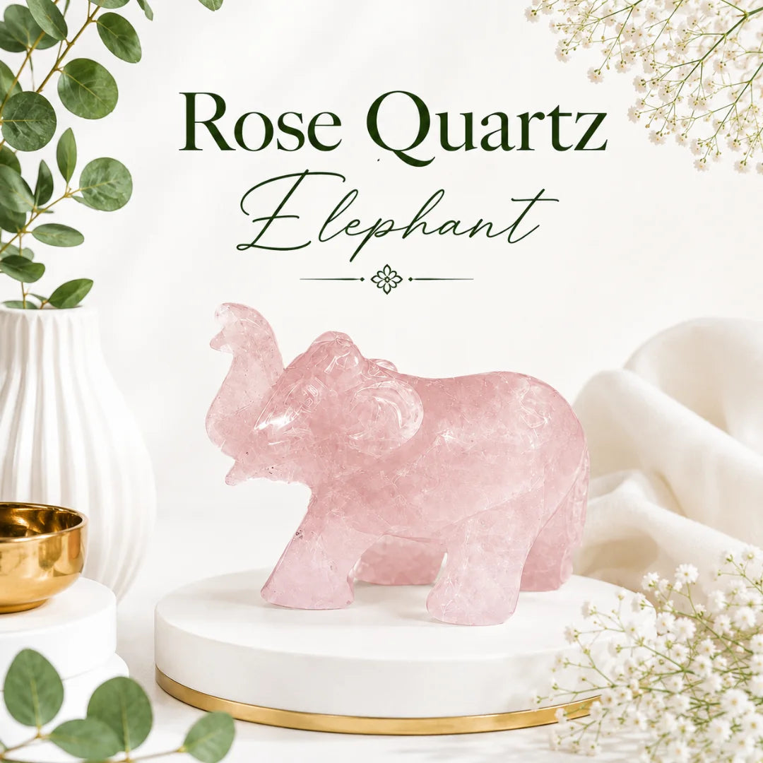 A pink rose quartz elephant figurine displayed on a white round pedestal with a gold base, surrounded by decorative greenery, white flowers, and soft fabric. Text at the top reads 'Rose Quartz Elephant'.