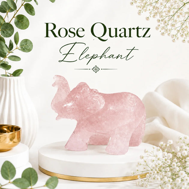 A pink rose quartz elephant figurine displayed on a white round pedestal with a gold base, surrounded by decorative greenery, white flowers, and soft fabric. Text at the top reads 'Rose Quartz Elephant'.