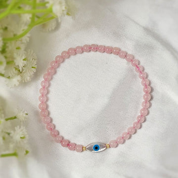 Pink beaded bracelet with a blue evil eye charm on a white fabric background with flowers.