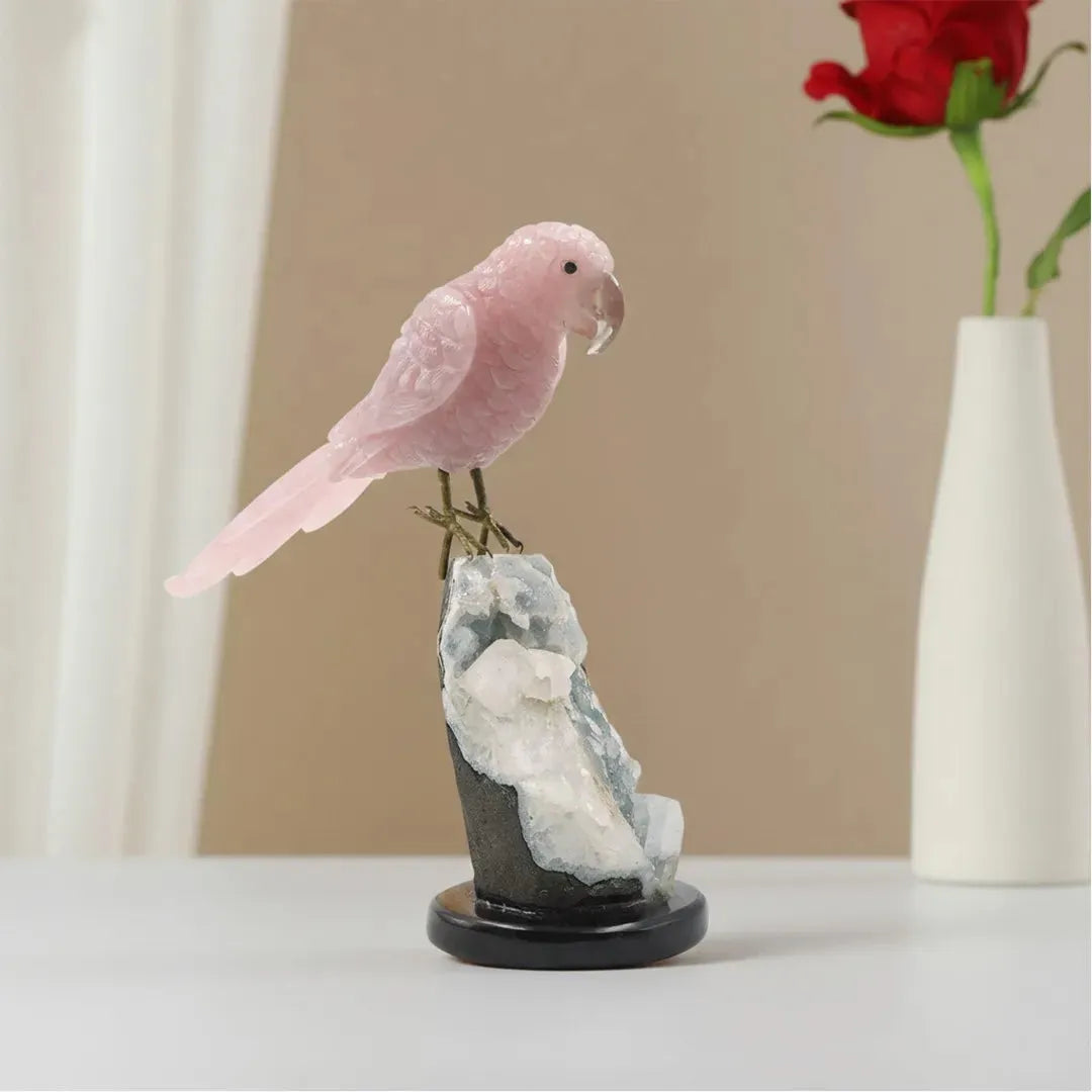 Pink parrot figurine on a rock with a red rose, against a beige wall.