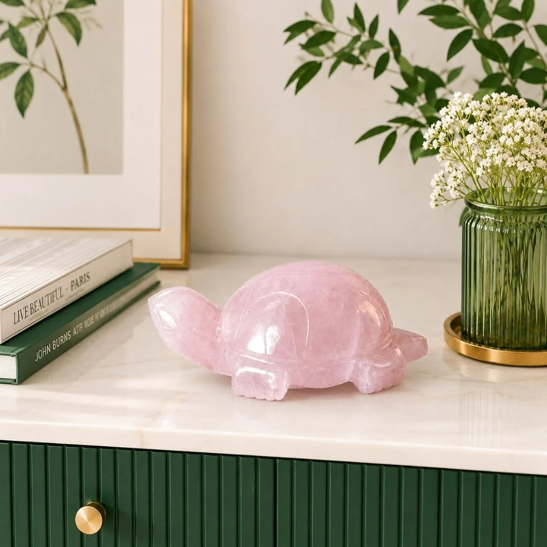 A polished pink rose quartz turtle figurine sits on a white marble countertop in a styled home setting, next to a green ribbed glass vase with baby's breath and a stack of books.