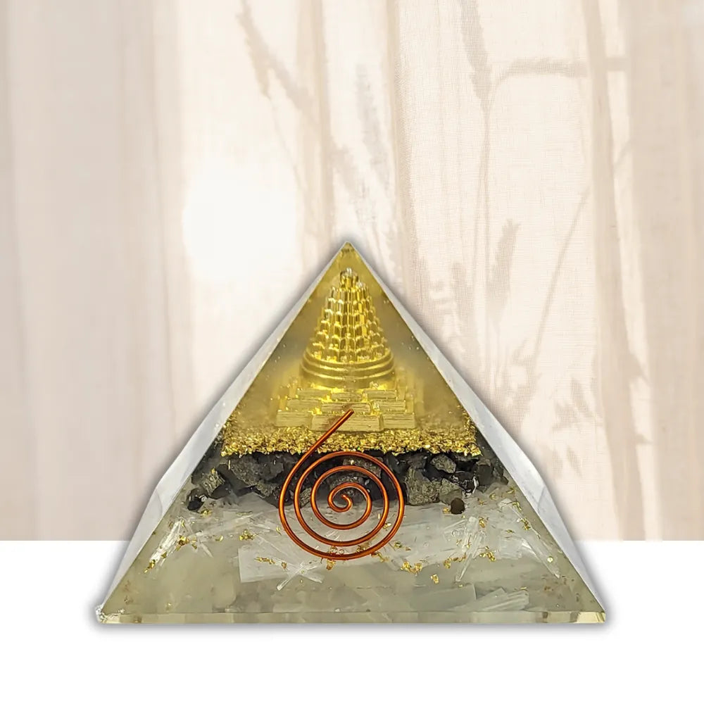 A triangular crystal pyramid or orichast stone with a gold-toned central structure, white quartz-like fragments, dark inclusions, and a copper spiral detail, shown against a soft beige backdrop with faint plant shadows.