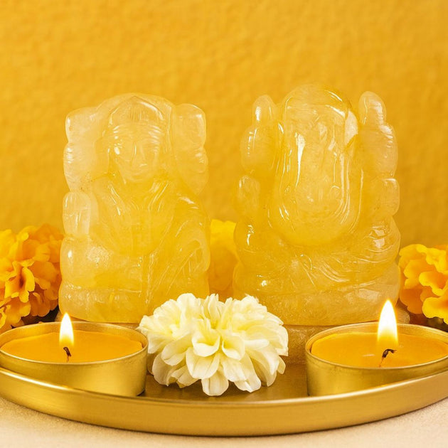 Two yellow aventurine crystal Lakshmi Ganesh on a golden colour tray with two lit candles and yellow flowers against a yellow background