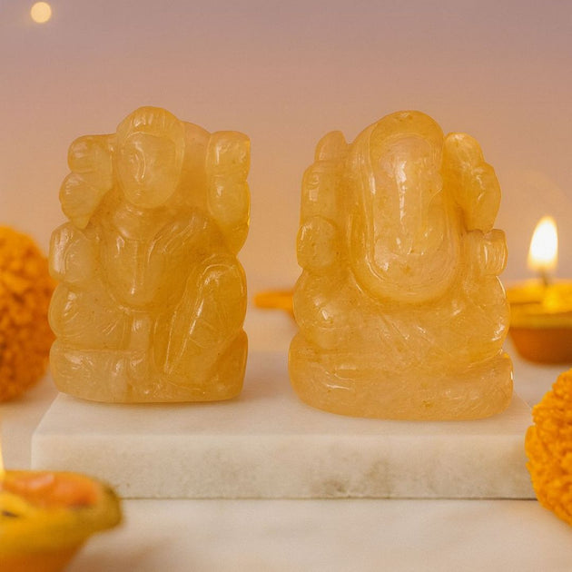 Two yellow stone statues of lakshmi ganesh on a marble base with a candle in the diya.