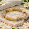 A beaded stretch bracelet made of round, polished gemstone-like beads in warm yellow, tan, cream, and light brown tones, displayed on a textured stone slab with white flowers and green leaves around it.
